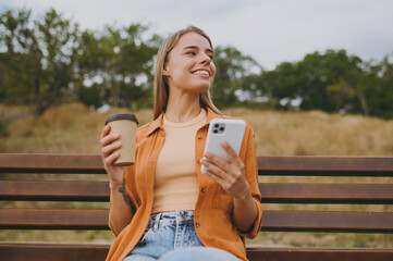 Young smiling woman wears orange shirt casual clothes use mobile cell phone drink coffee look aside...