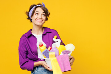 Young smiling happy woman wear purple shirt hold basin with detergent bottles do housework tidy up...