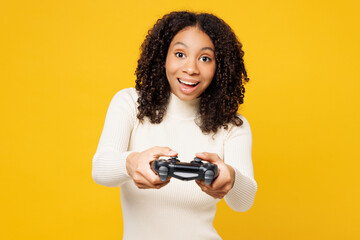 Little happy kid teen girl of African American ethnicity wear white casual clothes play pc game with joystick console isolated on plain yellow background studio portrait. Childhood lifestyle concept.