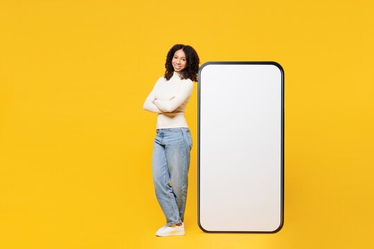 Full body fun little kid teen girl wear white casual clothes stand near big huge blank screen mobile cell phone smartphone with workspace area isolated on plain yellow background. Lifestyle concept.