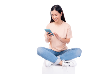 Relaxed young woman smiling while using her smartphone, sitting cross-legged on a peach background.
