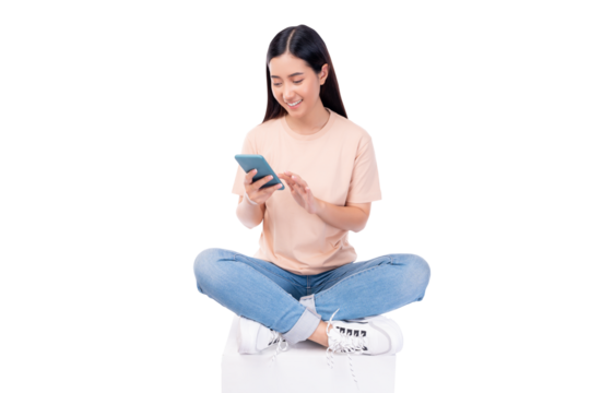 Relaxed young woman smiling while using her smartphone, sitting cross-legged on a peach background.
