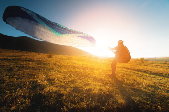 paraglider with a blue parachute takes off. A male athlete stands on the field and lifts a paraglider into the air