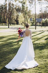 Wedding. The bride in a white dress stands on a green lawn in the forest and holds a bouquet of bright flowers in her hand