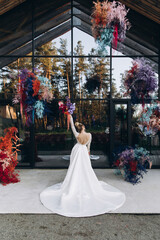 Wedding. The bride in a white dress stands against a glass wall and arrangements of colored flowers and greenery, holding a bouquet of bright flowers