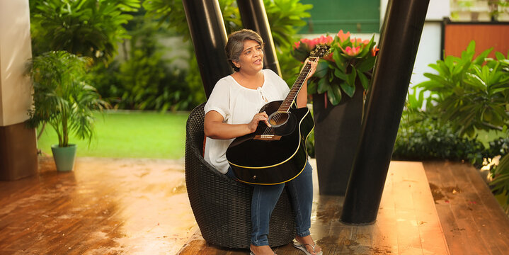 Asian Indian Hindu happy older gen x aged woman lady sitting chair singing song play guitar enjoy fun joy pastime outdoor home park smiling old elder adult female tuning music lyric house garden