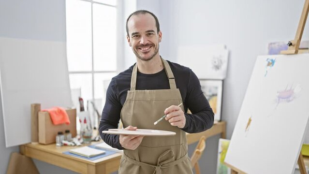 A confident bald man with a beard wearing an apron smiles while holding a paintbrush and palette in an art studio.