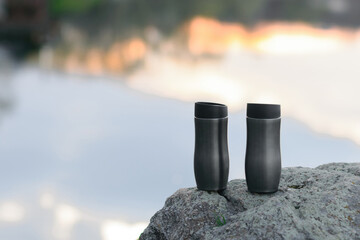 Tourist thermos mug on the background of mountains, stones and a river in which the sunset and...