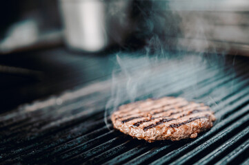 A sizzling burger patty cooks on a grill, smoke rising from its perfectly seared surface. Grill marks accentuate its juiciness.