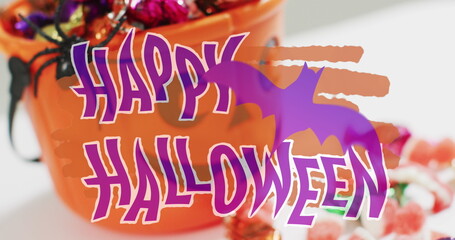 Happy halloween text banner with bat icon against pumpkin shaped bucket full of halloween candies