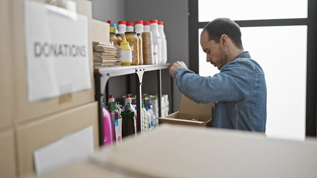 Hispanic man organizing donations in warehouse, wearing denim jacket and looking thoughtful amidst boxes and supplies.