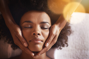 Woman, relax and massage on face with hands and care for facial, wellness and spa treatment on bed. Above, lens flare and african female person with skincare and cosmetics at hotel with skin glow
