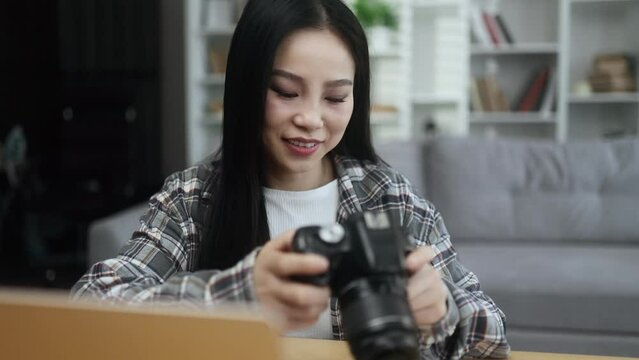 Portrait of pretty young asian woman photographer hold digital camera looking at screen choosing photos for editing while sitting in front of laptop computer at home workplace