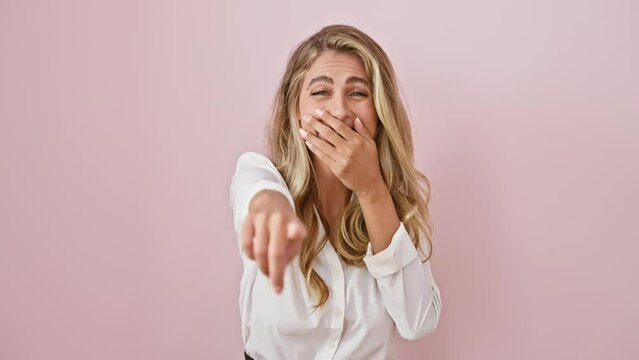 Joyful young blonde woman caught in a cheerful prank! standing in a pink isolated background, wearing a shirt, pointing finger at you, laughing with hand over mouth. can you match her happiness?