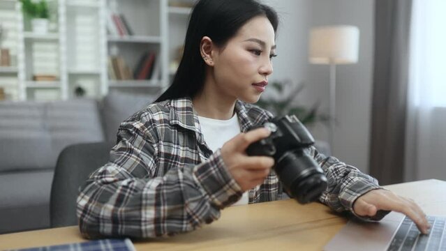 Portrait of pretty young asian woman photographer hold digital camera looking at screen choosing photos for editing while sitting in front of laptop computer at home workplace