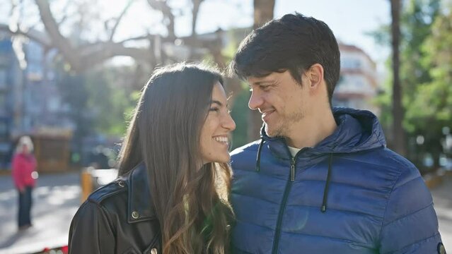 Smiling couple in love standing together outdoors in an urban park, with trees and sunlight in the background.