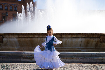 A pretty little girl dancing flamenco dressed in a white dress with ruffles and blue fringes in a...