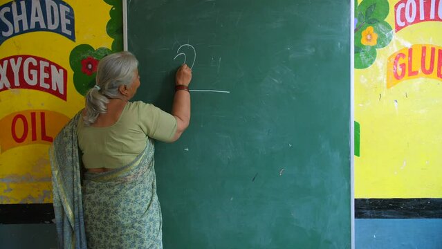 Grey-haired Indian teacher teaching school kids how to draw on blackboard - drawing class  government school. Old age teacher planning fun activity for students during free period in school - creat...