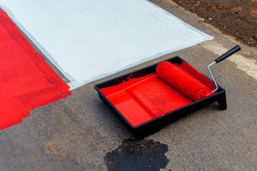 Paint roller in tray with red paint on asphalt road, painting a pedestrian crossing.