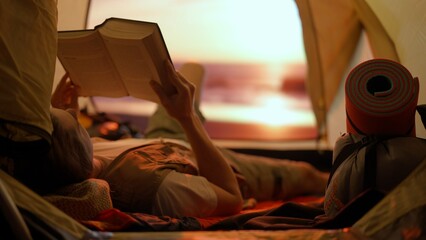 Person on campsite traveling and hiking, exploring nature. Young man laying reading a book inside the tent at sunset on the beach, holiday and vacation outdoors. - obrazy, fototapety, plakaty