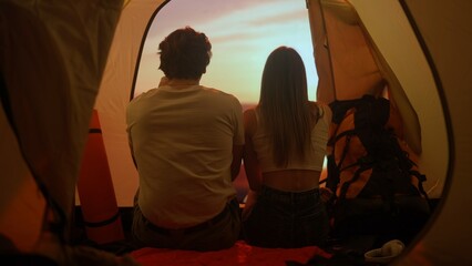 People on campsite traveling and hiking, exploring nature. Young loving couple sitting inside the tent looking at sunset on the beach, holiday and vacation outdoors.