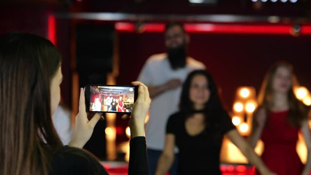 Back view of woman holding mobile phone and making video of her friends dancing in the nightclub. Lifestyle, dance, disco concept. Slow motion