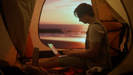 Person on campsite traveling and hiking, exploring nature. Young man sitting inside the tent at sunset on the beach working on laptop, holiday and vacation outdoors.