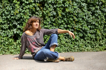 A young model beautiful woman girl in denim trousers stands near a wall overgrown with climbing plants. Background with plant leaves.