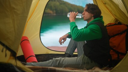Person on campsite traveling and hiking, exploring nature. Young man sitting drinking water inside the tent at lake coast in mountains, holiday and vacation outdoors.