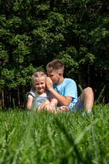 Fototapeta na wymiar The boy in casual attire leans close, whispering to the girl in denim, a tender sibling scene set against the park's verdant backdrop, National siblings day and happy friendships.