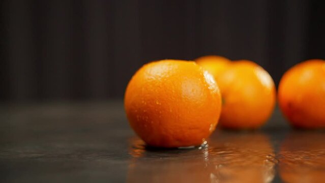 A group of oranges are featured in a video where they are being splashed with water and falling into water on a black surface. The oranges are also shown sitting on a table with water drops