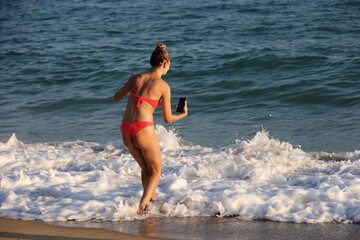 Girl in red bikini taking photos on smartphone camera of surf waves standing on sea beach