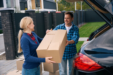 Joyful couple standing and unloading boxes out of car trunk