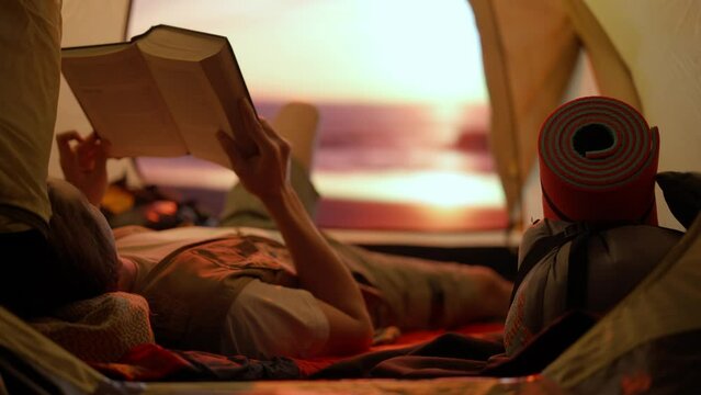 Person on campsite traveling and hiking, exploring nature. Young man laying reading a book inside the tent at sunset on the beach, holiday and vacation outdoors.