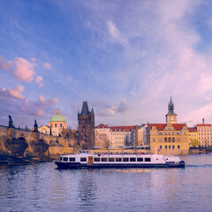 A boat glides on the water in front of a grand castle, surrounded by spring flowers along the Prague River.