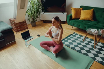 Rolgordijnen Peaceful woman is sitting in a lotus position on a yoga mat in a living room © Daria