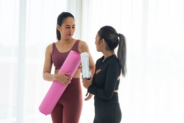 Two women in athletic wear engaging in conversation, one holding a yoga mat and the other a water...