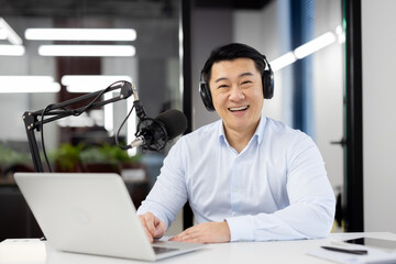 A smiling young Asian man is working in the office, sitting at a desk in headphones, and conducting...
