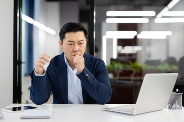 Young Asian male businessman sitting at desk in office and suffering from severe cough and asthma...