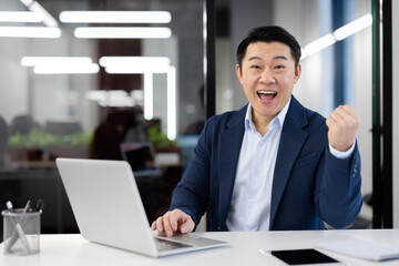 Portrait of a happy young Asian man in a business suit sitting in the office at the table in front of a laptop and looking at the camera with success and achievement, showing a victory with his hand.