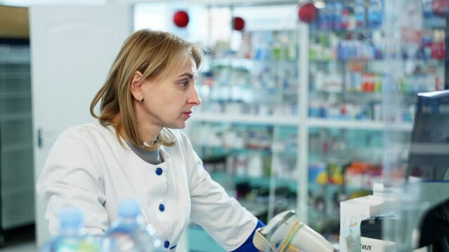 A dedicated female pharmacist in a white coat is seen using a computer to assist with her tasks in a clinical pharmacy setting, showcasing the daily responsibilities of a health care scientist