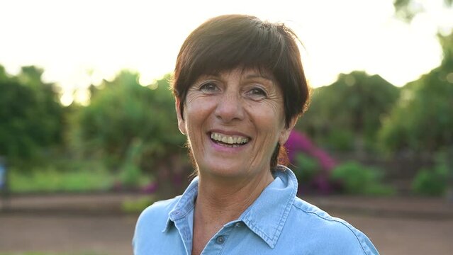 Senior caucasian woman smiling on camera at city park with sunrise in background - Joyful elderly and pension concept