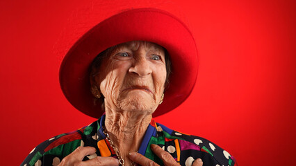 Closeup funny view of happy elderly toothless senior woman wearing red hat pointing fingers herself ask say who me, smiling talking and pointing. Isolated on red background.