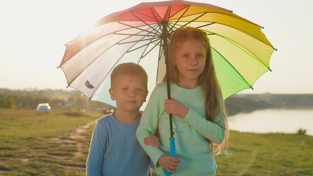 Siblings huddle beneath colorful umbrella