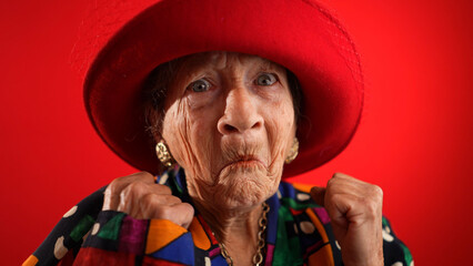 Saying WOW, a happy fisheye closeup portrait caricature of funny elderly woman with red hat isolated on red background.