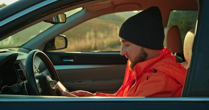 A young man dressed in an orange jacket and cap, in a car with a phone in his hands.