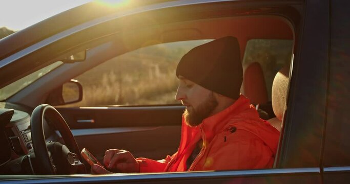 A young man dressed in an orange jacket and cap, in a car with a phone in his hands.