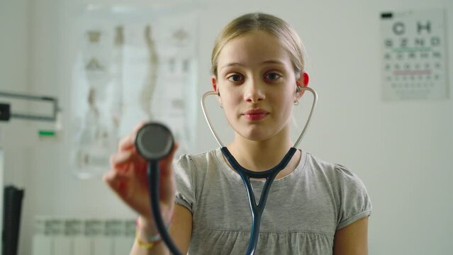 Portrait of a little girl in a medical clinic, wearing stethoscope and showing it at camera.