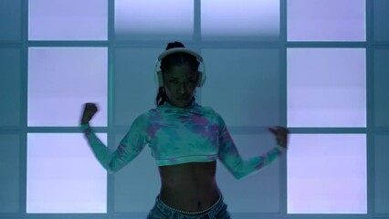 A beautiful Latin girl, African American, as soon as she puts on her headphones the lights come on and she starts dancing to hip hop music in front of a neon blue background.