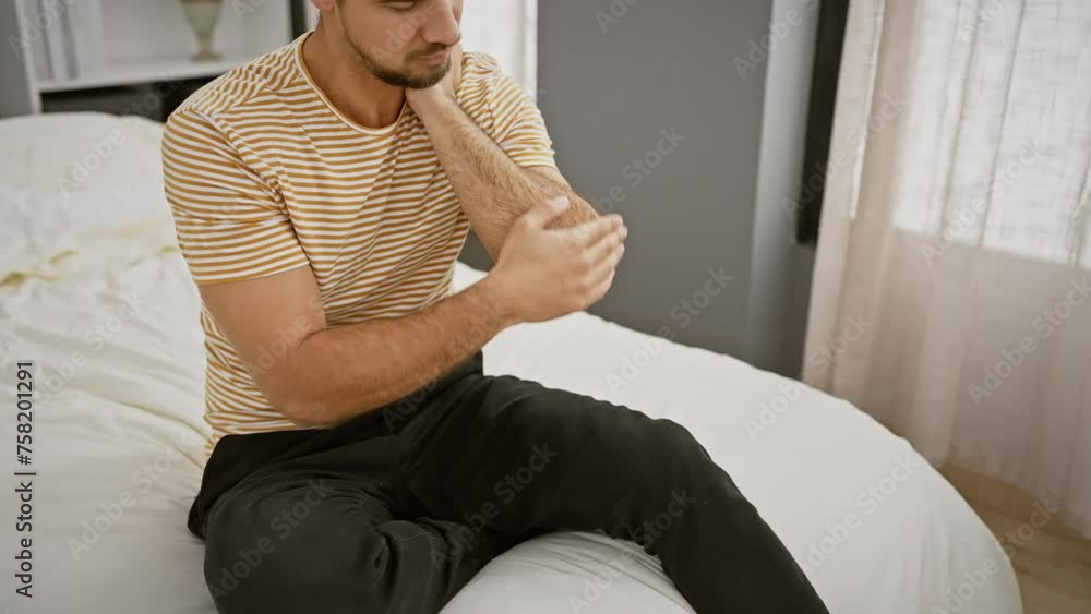 Wall mural Hispanic man applying lotion to elbow in a cozy bedroom setting, conveying a personal care routine.